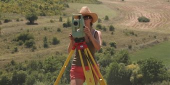 A person stands in a field wearing sunglasses and a sun hat, measuring the field.