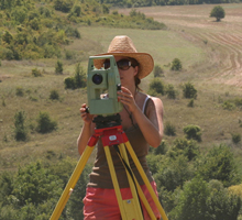 A person in a field, wearing sunglasses and a sunhat, measures the land