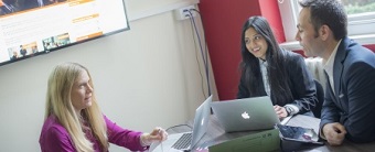 Photo of two women and a man sat in discussion at a table with a selection of laptops and books open in front of them.