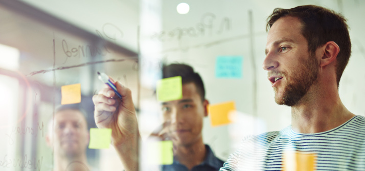 Three people standing at a clear messaging board with writing and post it notes on it